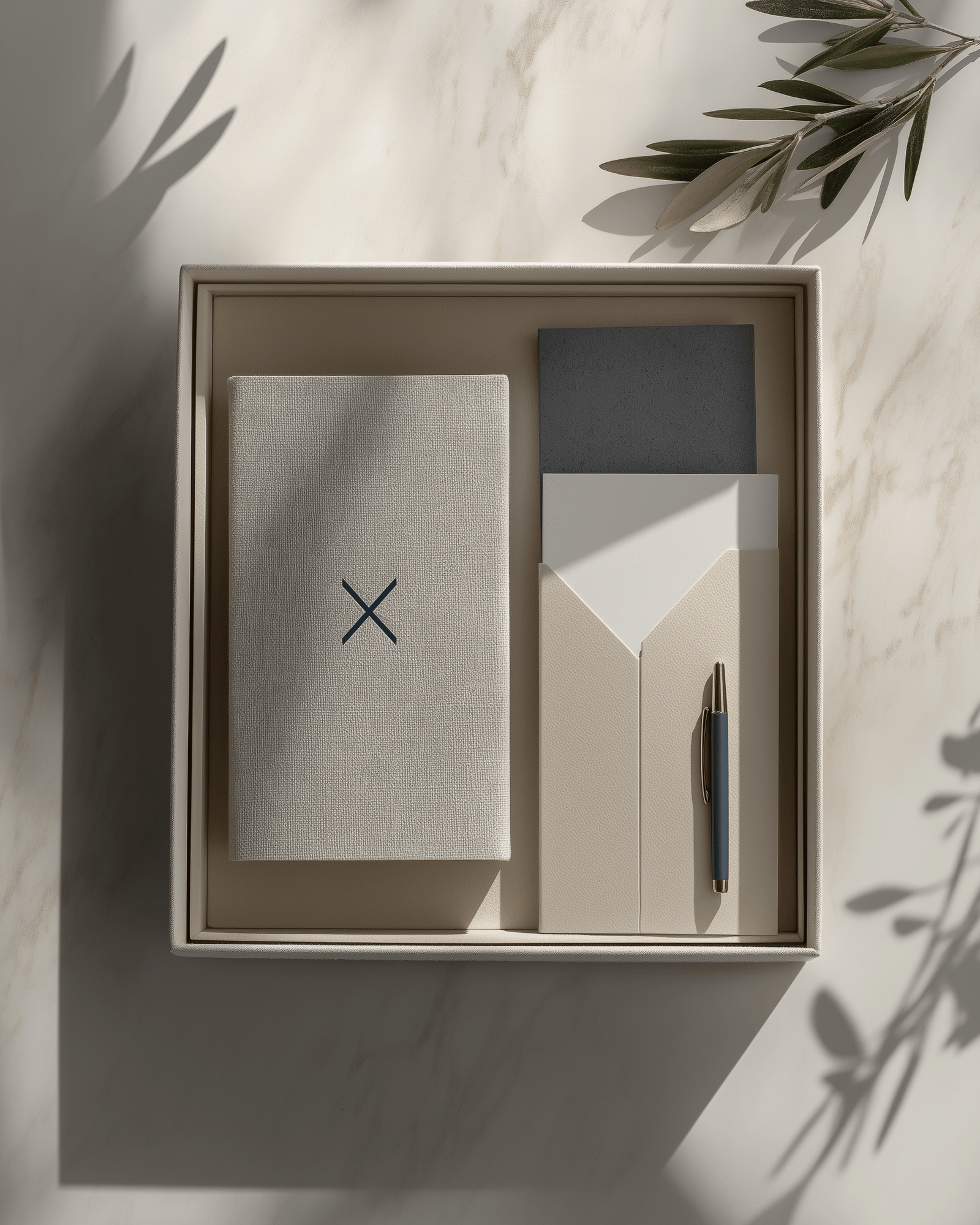 An overhead photograph of an editorial presentation box: a linen-bound book with a navy mark on the cover, a folded letter, a slate notebook, and a navy-and-brass pen, framed by olive branches in soft afternoon light.